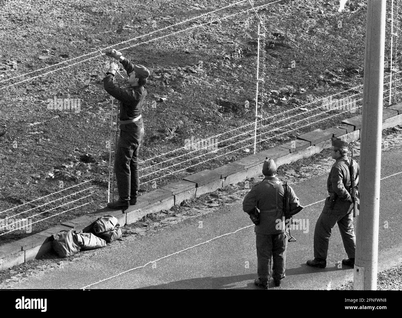 Berlin-Neukoelln / GDR / Wall / 10 / 1980 le guardie di frontiera della GDR tirano il nuovo filo spinato lungo il muro, Bouchestrasse tra Neukoelln e Treptow. È carica elettricamente e attiva un allarme quando viene toccato. In alto a sinistra si trova Berlino Ovest. Il muro è dipinto di bianco per far rischiare i rifugiati. Le due guardie di confine davanti fanno attenzione che le altre non scappino in Occidente. // fili dentati / fortificazioni di frontiera / distretti [traduzione automatizzata] Foto Stock