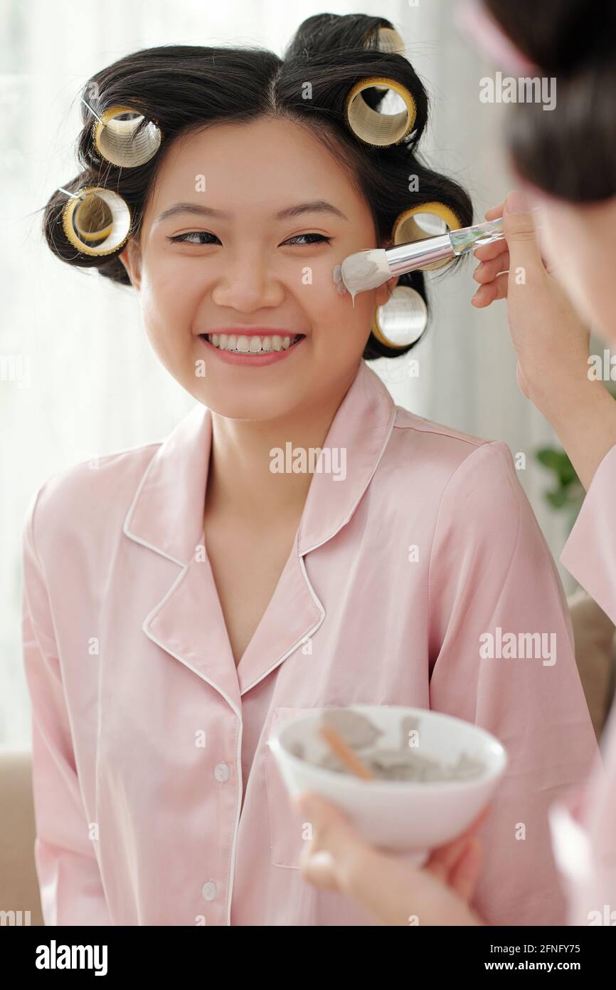 Ritratto di giovane donna asiatica graziosa con i rulli dei capelli che ottengono maschera di argilla applicata sulla sua faccia Foto Stock