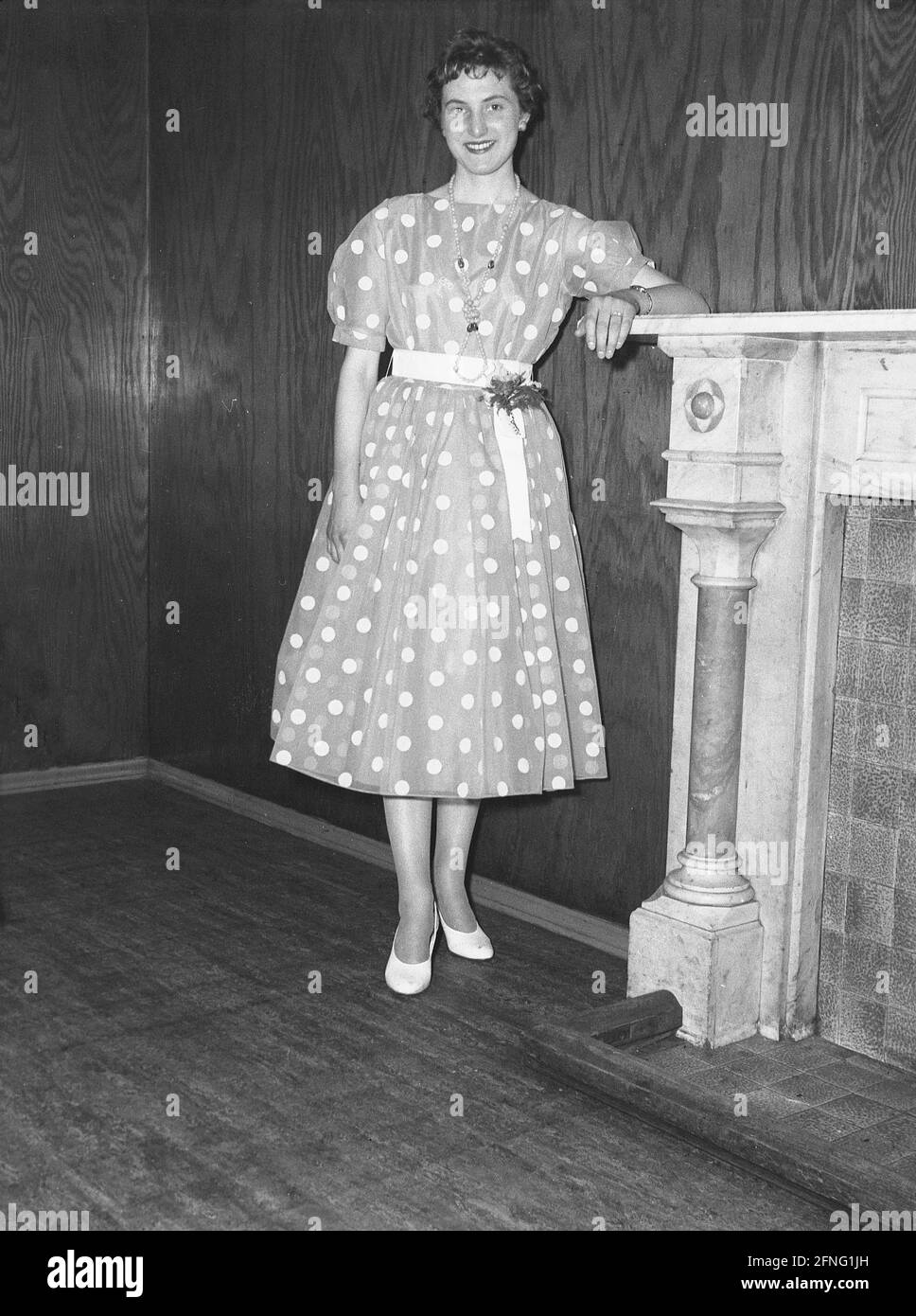 anni '50, storico, una giovane donna in piedi accanto a un camino nella sua festa di schiumata, un poka-dot o vestito macchiato, un capo di abbigliamento in moda alla fine degli anni '50, quando rockabiliy e la nascita di rock e roll stava per accadere. Foto Stock