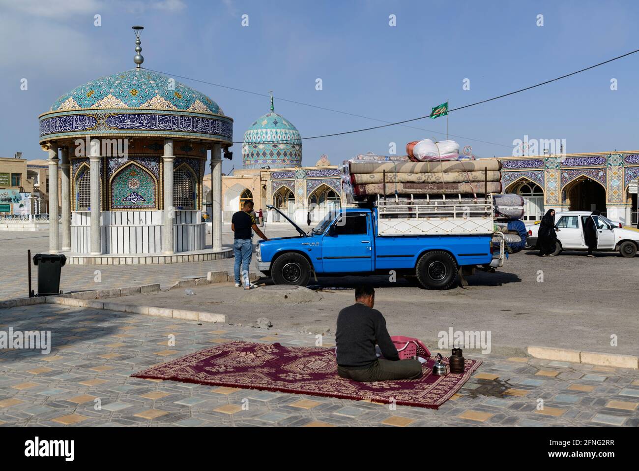 Pick-up camion blu caricato con tappeti persiani e aperto il cofano dietro un uomo seduto su un tappeto vicino a una moschea di Nain, Contea di Nain, Provincia di Isfahan, i Foto Stock