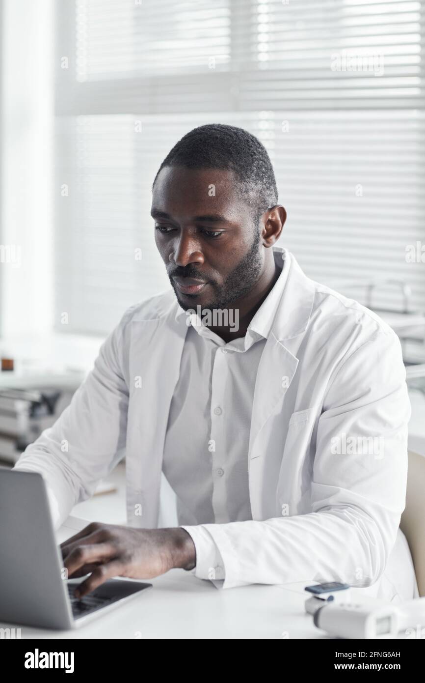 Giovane medico africano in camice bianco seduto alla scrivania e lavorare su un computer portatile in ufficio Foto Stock