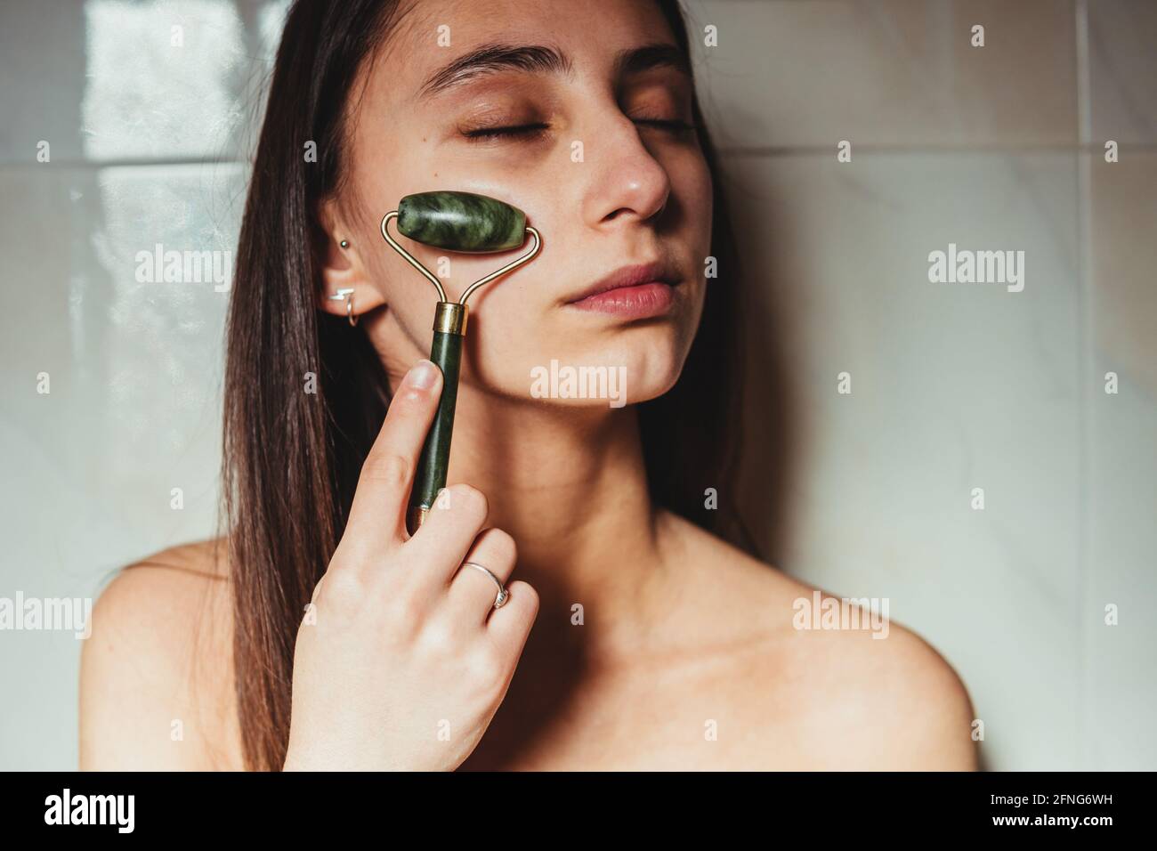 Raccolto concentrato giovane femmina con occhi chiusi massaggiando guancia con rullo jade contro parete in ceramica Foto Stock