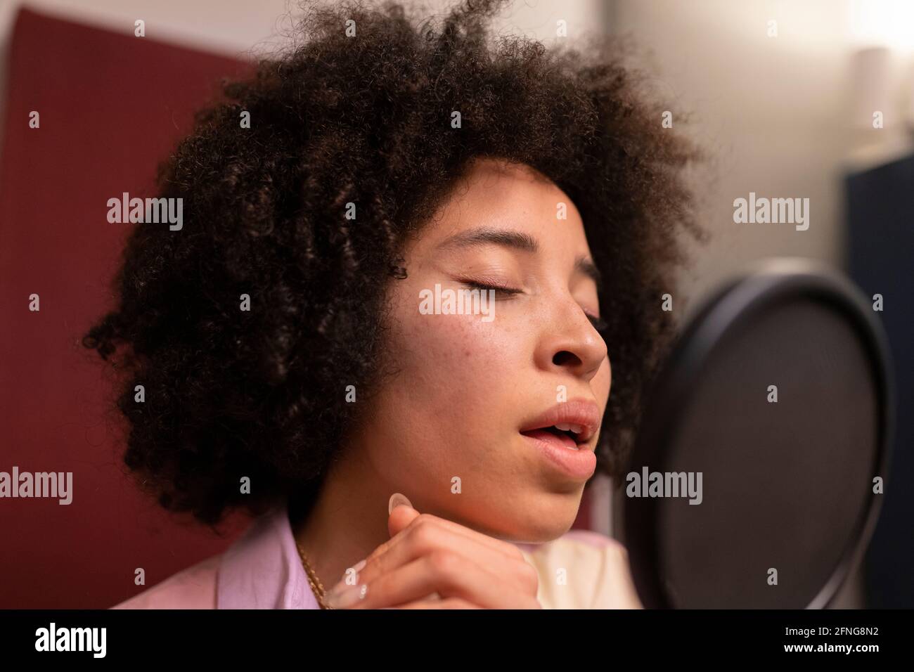 Cantante femminile nera che esegue canzoni contro il microfono con filtro pop mentre gli occhi sono in piedi e chiusi sullo studio audio Foto Stock