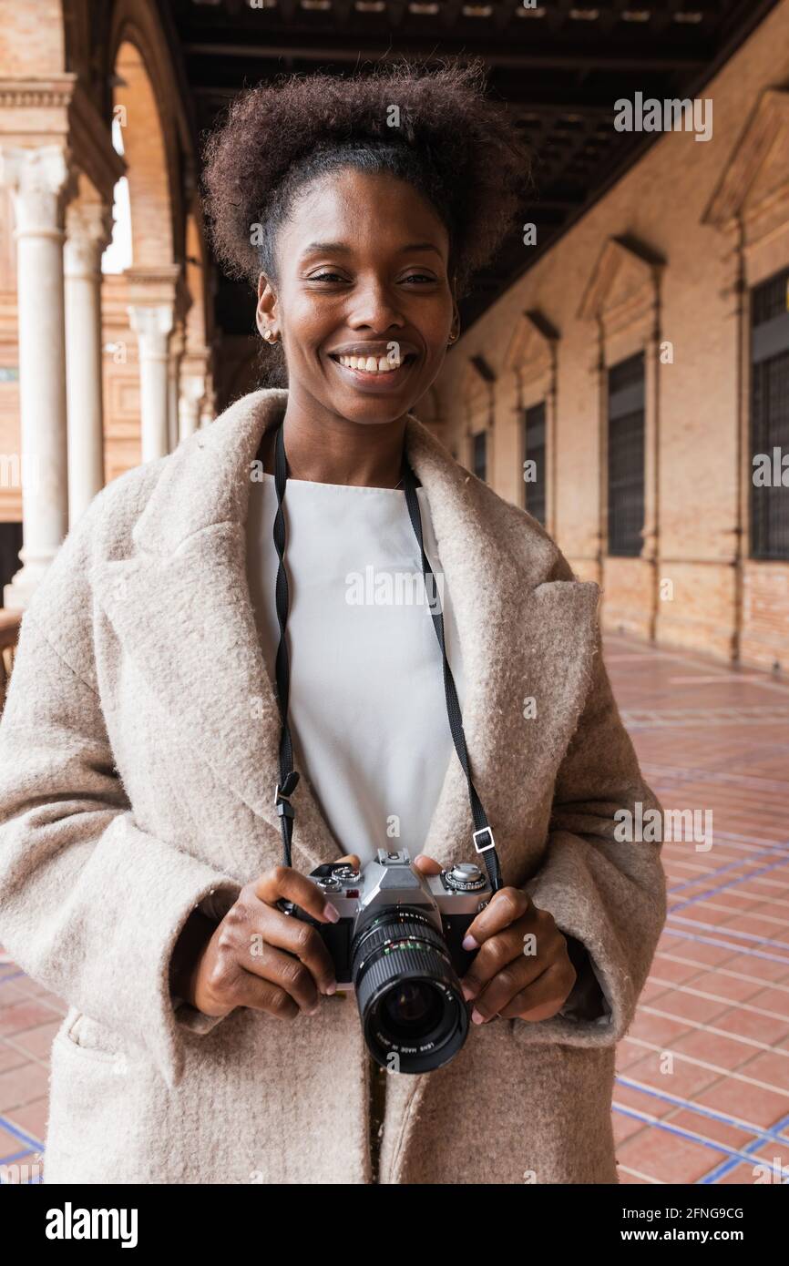 Vista frontale della giovane fotografa afroamericana in caldo cappotto in edifici storici della città con una moderna fotocamera primo giorno di primavera Foto Stock