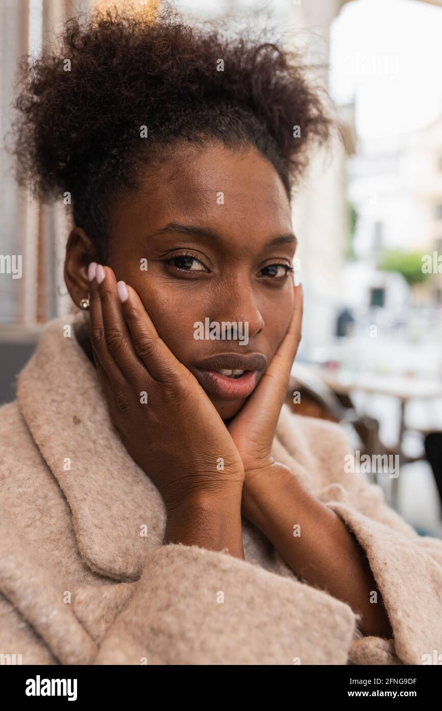 Positiva giovane afroamericana femmina in caldo cappotto che tocca le guance e guardando la macchina fotografica sulla strada soleggiata della città Foto Stock