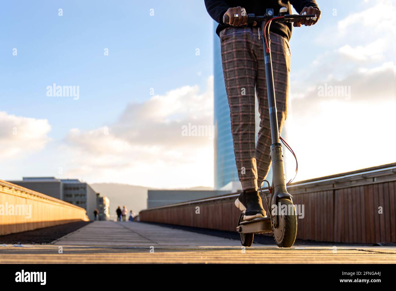 Un imprenditore etnico maschile non riconoscibile in sella a uno scooter elettrico in città ponte passerella contro gli edifici sotto il cielo blu nuvoloso Foto Stock