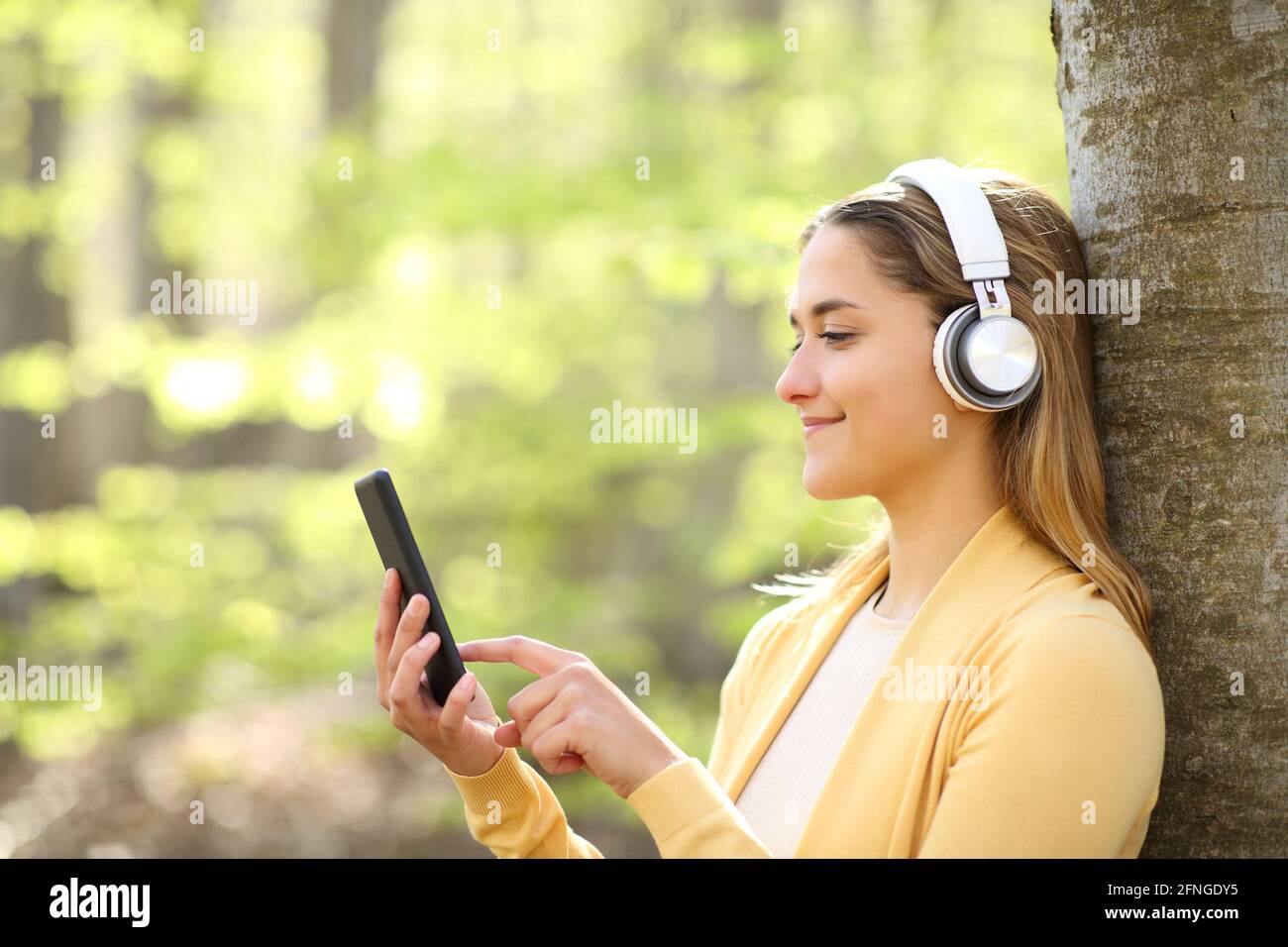 Ritratto in vista laterale di una donna felice che indossa le cuffie alla musica utilizzando lo smartphone in una foresta Foto Stock