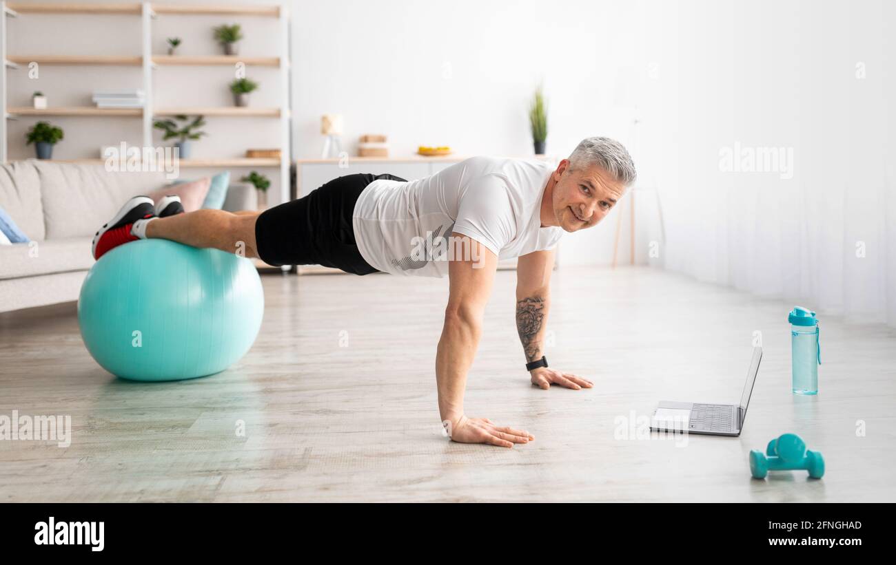 Formazione online nazionale. Uomo maturo in piedi in tavola sulla palla di fitness, allenarsi e guardare video tutorial sportivi Foto Stock