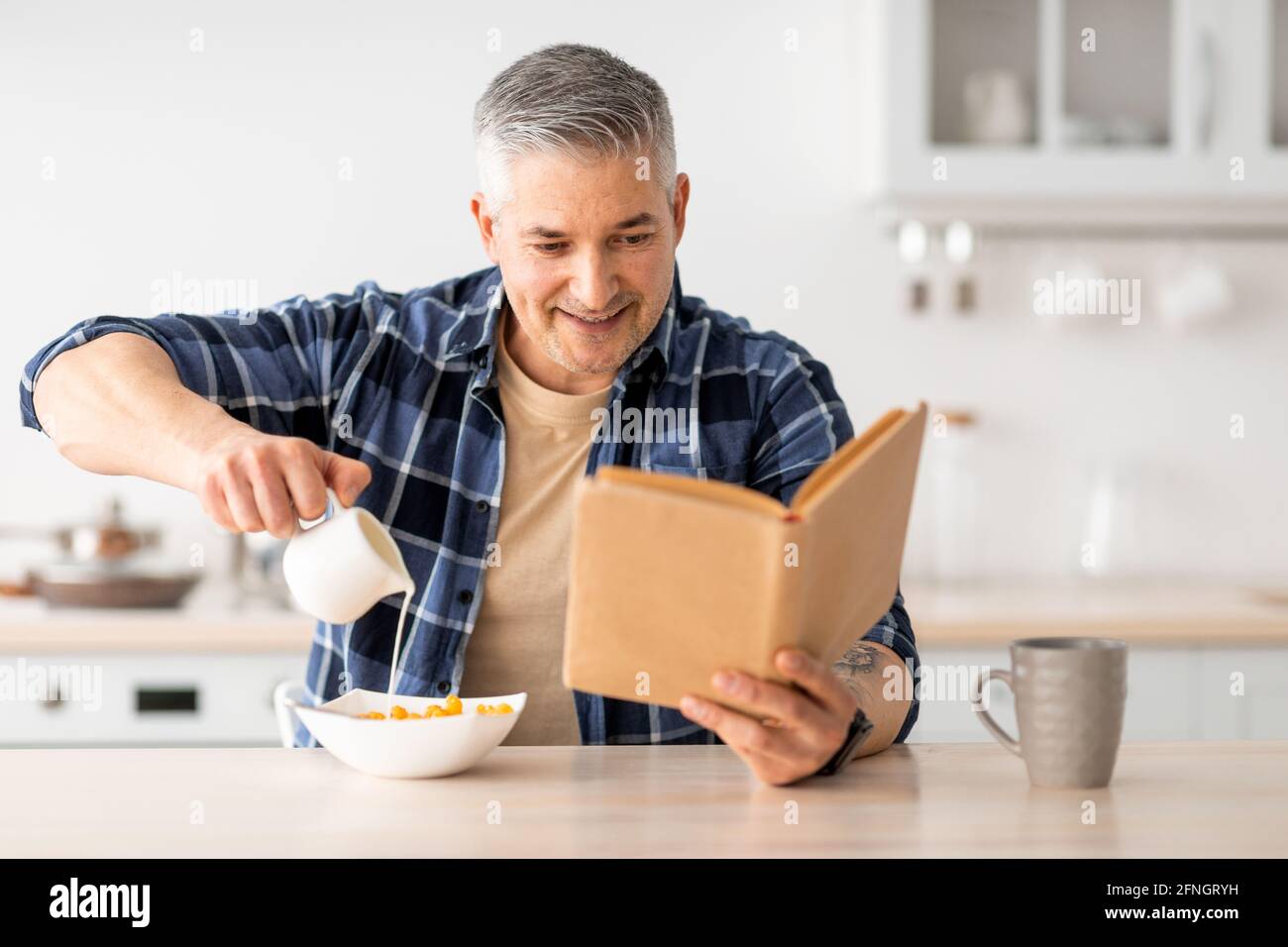 Felice uomo anziano leggere libro e poring latte in cereali, facendo colazione in cucina interno Foto Stock