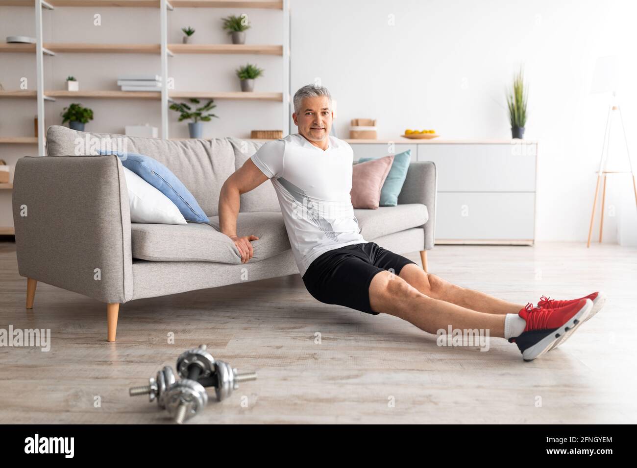 Allenamento a casa per mantenersi in forma. Felice uomo maturo appoggiato sul divano e facendo esercizi, allenandosi all'interno del soggiorno Foto Stock