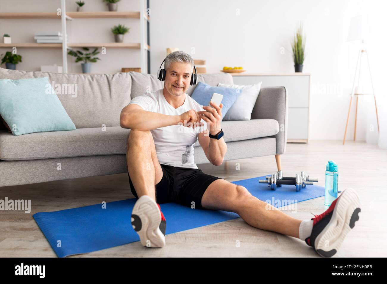 Musica per l'allenamento. Felice uomo di mezza età con le cuffie seduti sul tappetino e utilizzando lo smartphone, allenandosi a casa Foto Stock