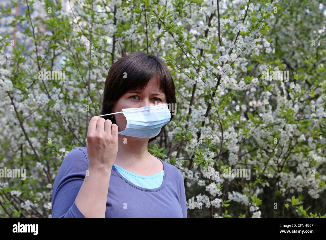 Donna che rimuove maschera medica viso in un giardino di primavera su fondo fiorito di ciliegio. Concetto di godere l'odore dei fiori, fine della quarantena Foto Stock