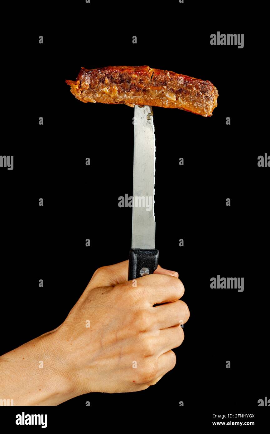 Immagine concettuale per il consumo di carne una persona sta tenendo un coltello bistecca in posizione verticale con una fetta di carne di manzo o di maiale inserita nella parte superiore. Isolamento versatile Foto Stock