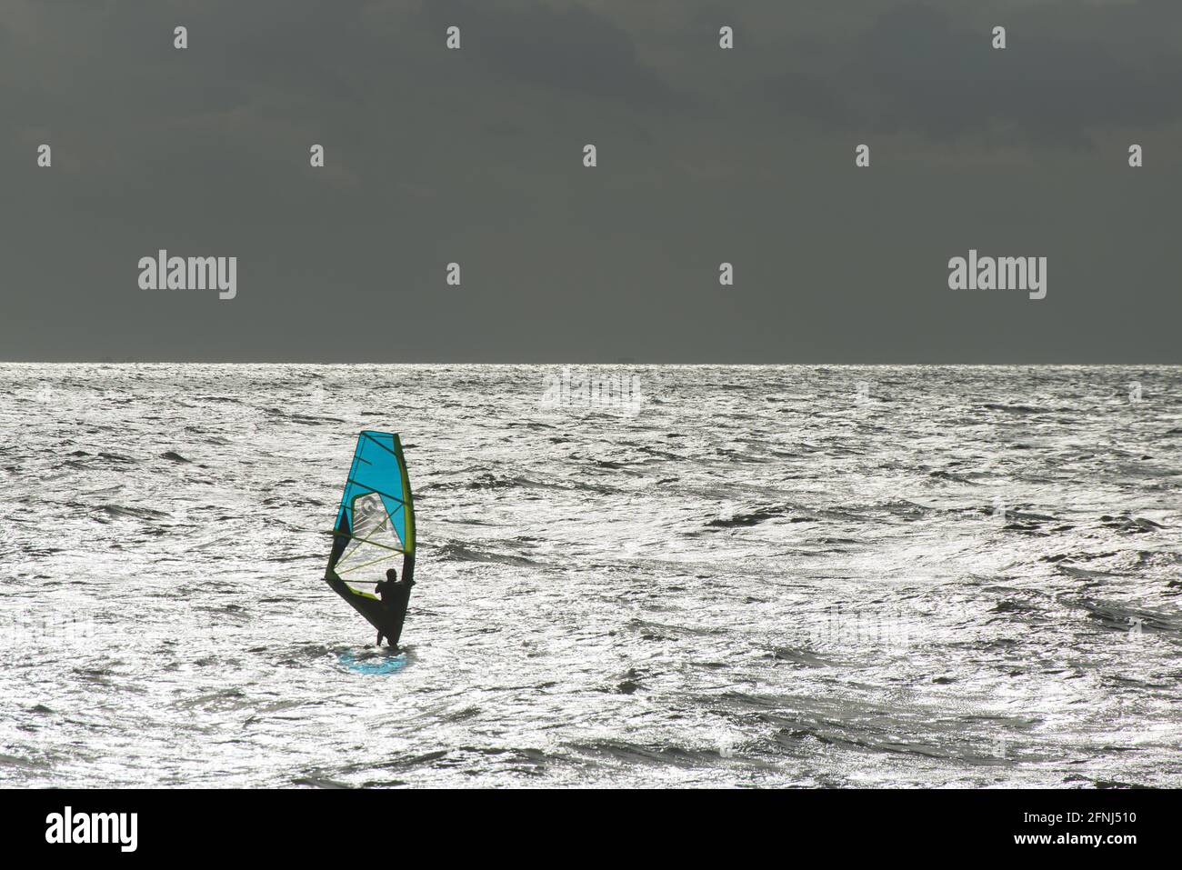 Immagine memorabile di un singolo windsurf con vela blu un mare argentato con orizzonte argentato che separa lo sfondo del cielo scuro Foto Stock