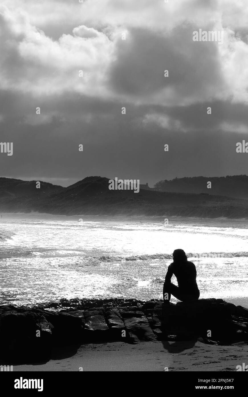 Un'immagine monocromatica in formato verticale di una singola figura in silhouette seduta su rocce in un ambiente costiero con sfondo di dune e nuvole in deriva Foto Stock