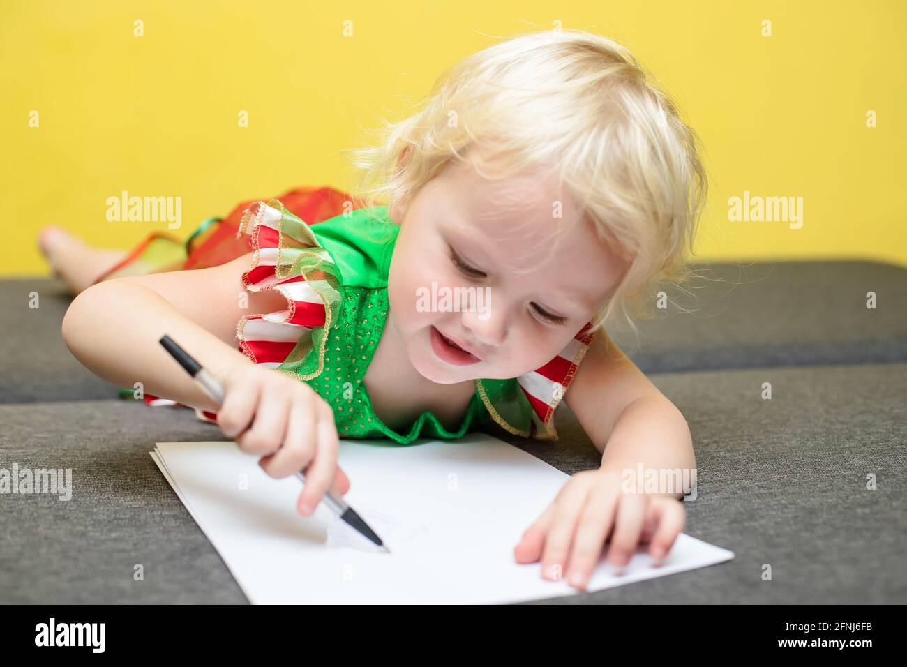 Ragazza scrive una lettera a Babbo Natale. Un bambino in costume Elf giace contro il muro giallo e fa auguri per Natale. Concetto di nuovo anno 2022. Foto Stock