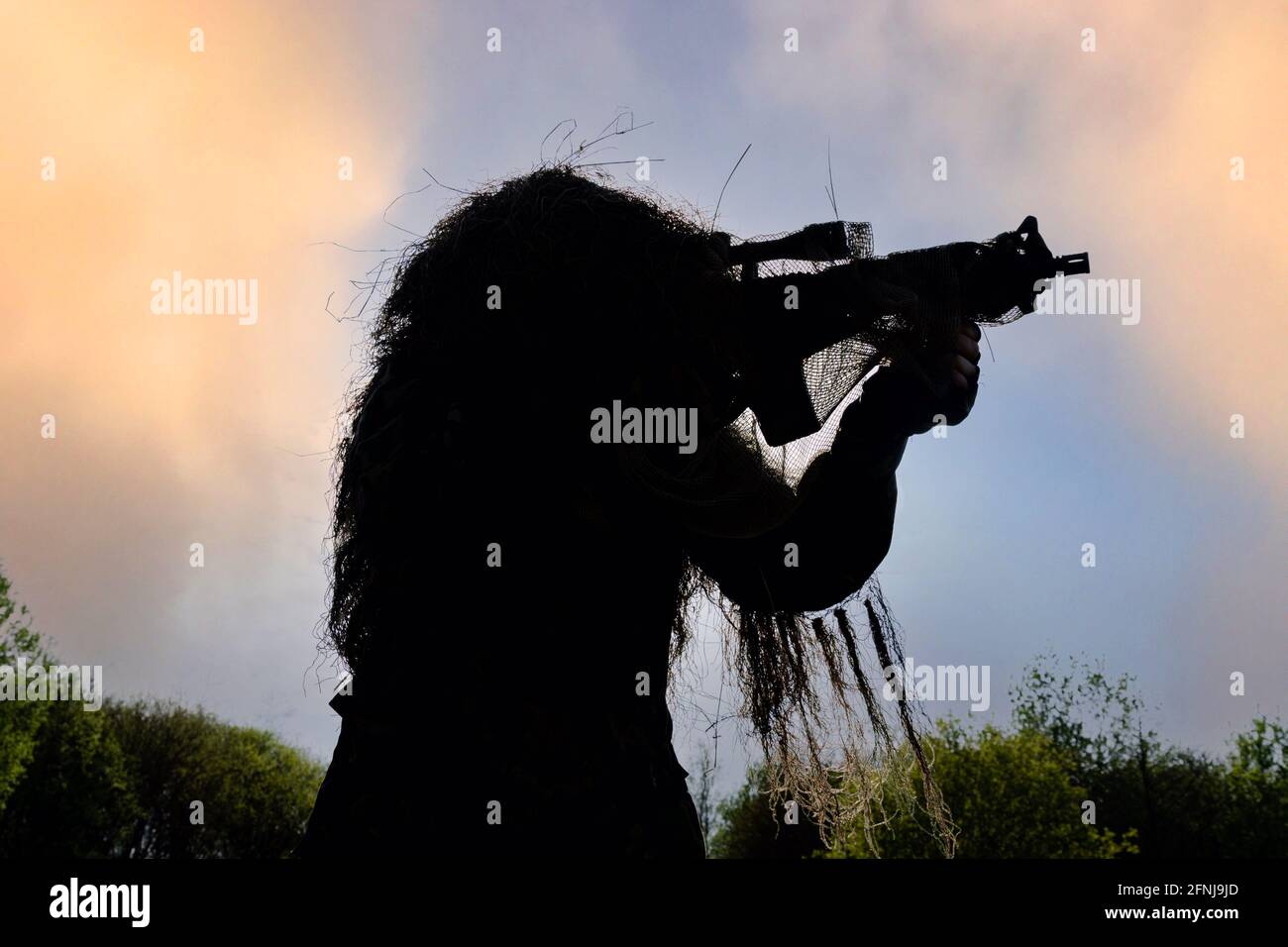 Sniper markman in completo Ghillie tuta in piedi e di puntamento Silhouette contro il cielo con la foresta che viene visualizzata nel Sfondo Foto Stock