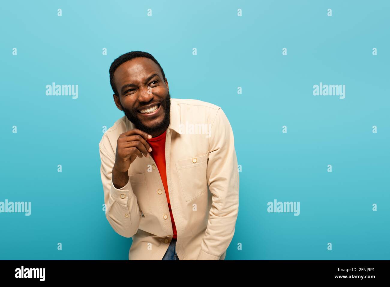 uomo afro-americano malizioso sorridente mentre si guarda lontano isolato sopra blu Foto Stock