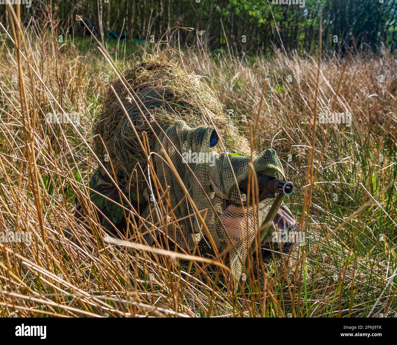 Sniper in una tuta Ghillie camuffata nel lungo Grass Mira a un obiettivo nemico attraverso l'ambito Foto Stock