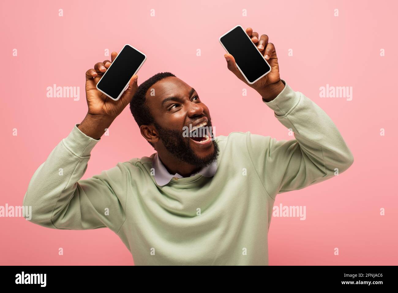 Uomo afroamericano stupito che guarda i telefoni cellulari isolati su rosa Foto Stock