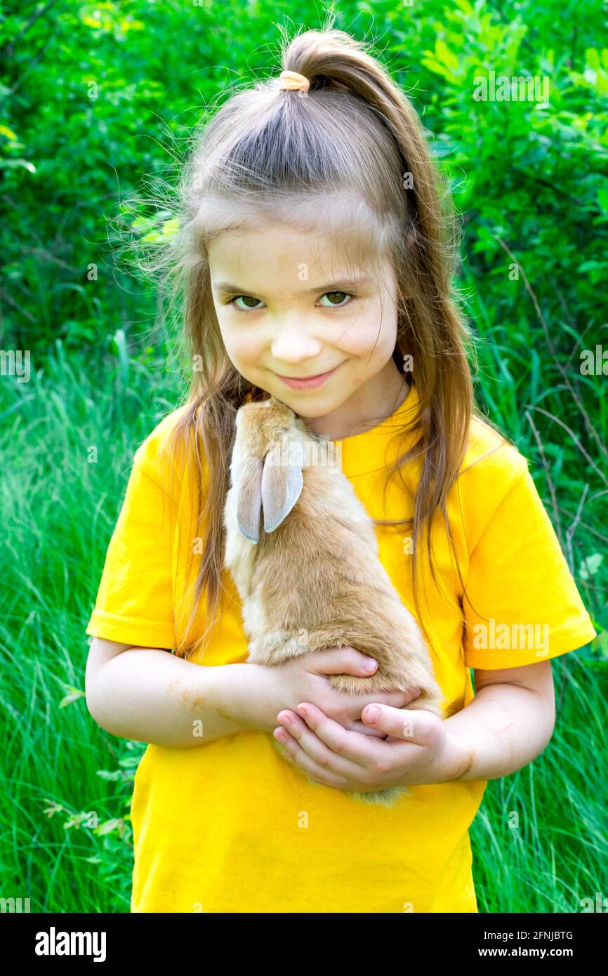 La ragazza carina piccola gioca con un coniglio di zenzero reale su uno sfondo di piante verdi. Attività estive all'aperto per bambini con animali domestici. Ragazza che gioca con Foto Stock