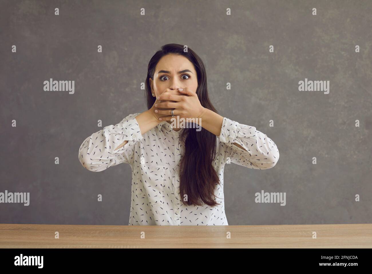 Giovane donna spaventa coprendo la bocca con la mano seduta al tavolo scatto in studio Foto Stock