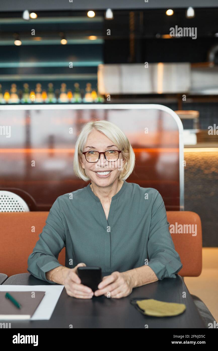 Ritratto di allegra donna d'affari caucasica con capelli biondi in occhiali con smartphone e sorridendo a macchina fotografica in un caffè moderno Foto Stock