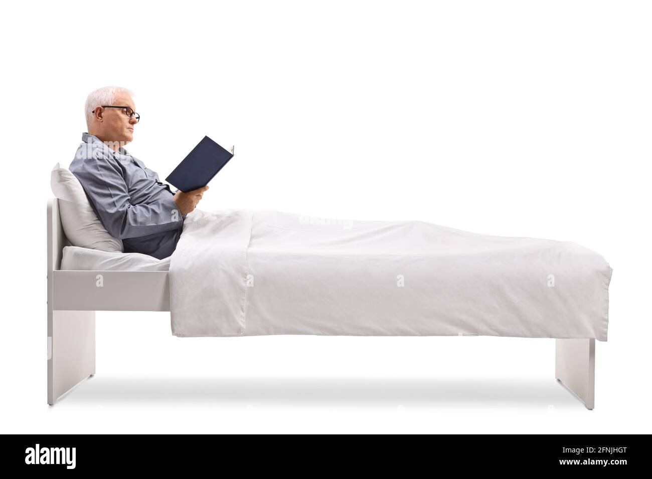 Uomo maturo in pigiama seduto su un letto sotto un piumone e leggere un libro isolato su sfondo bianco Foto Stock