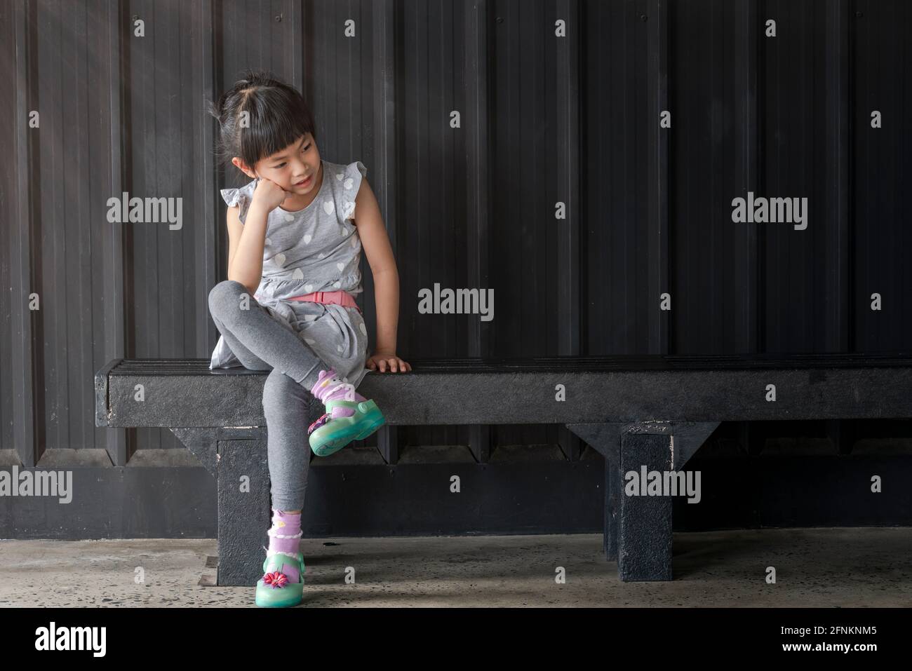 Ritratto Asian bambina ragazza seduta su metallo panca nero, sfondo nero. Siediti con le gambe incrociate, il gomito sul ginocchio e il viso sulla mano. La ragazza Foto Stock