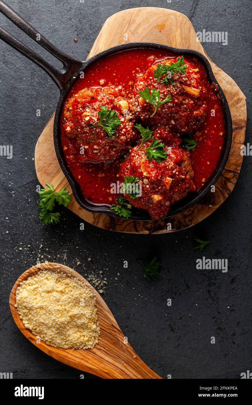 Vista ravvicinata di tre polpette tradizionali italiane di grandi dimensioni in salsa di pomodoro sfrigolante in padella di ghisa. La padella si siede su trivet di legno e foglie di prezzemolo Foto Stock