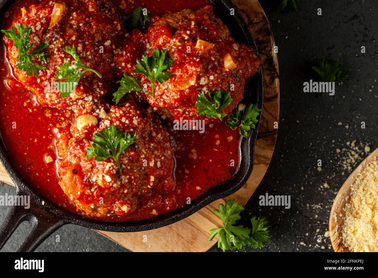 Vista ravvicinata di tre polpette tradizionali italiane di grandi dimensioni in salsa di pomodoro sfrigolante in padella di ghisa. La padella si siede su trivet di legno e foglie di prezzemolo Foto Stock