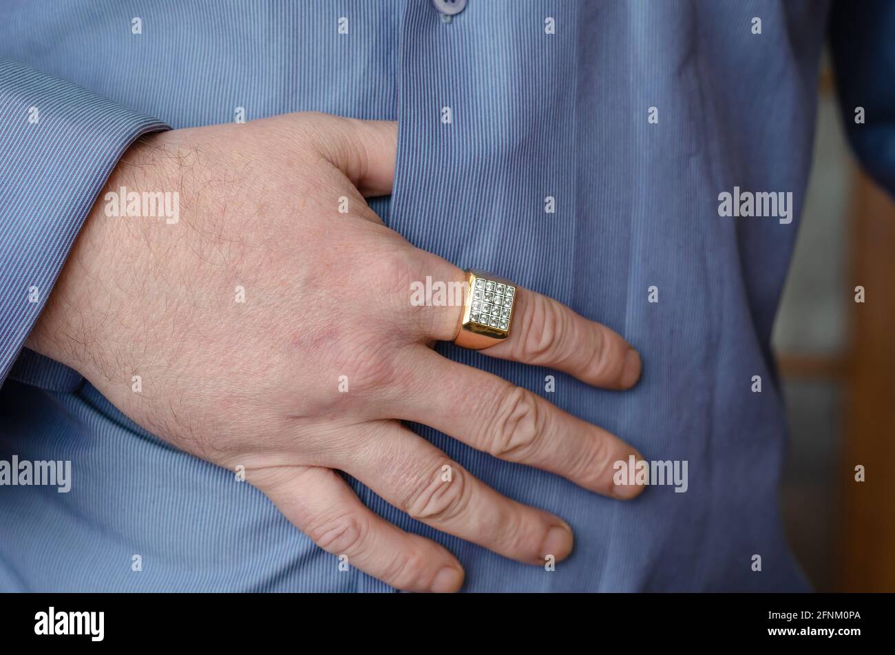 Un primo piano di una mano con un anello d'oro sul dito indice. Un uomo di  mezza età sta tenendo la mano su una camicia blu nella parte centrale del  suo corpo