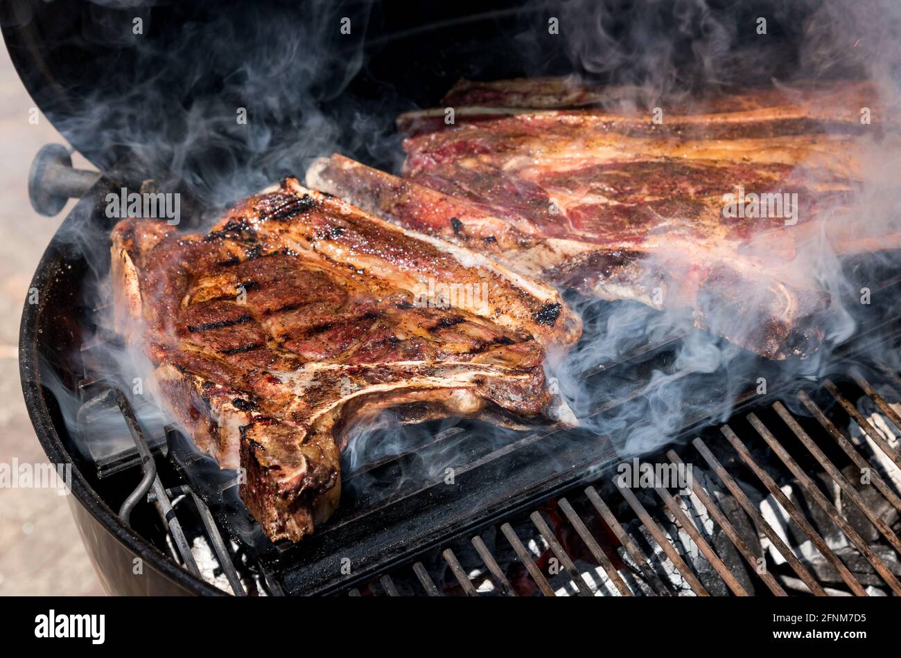 Bistecche di manzo stagionate non disossate grigliate su un fuoco al barbecue frizzante sopra i carboni caldi con fumo in un primo piano vista esterna Foto Stock