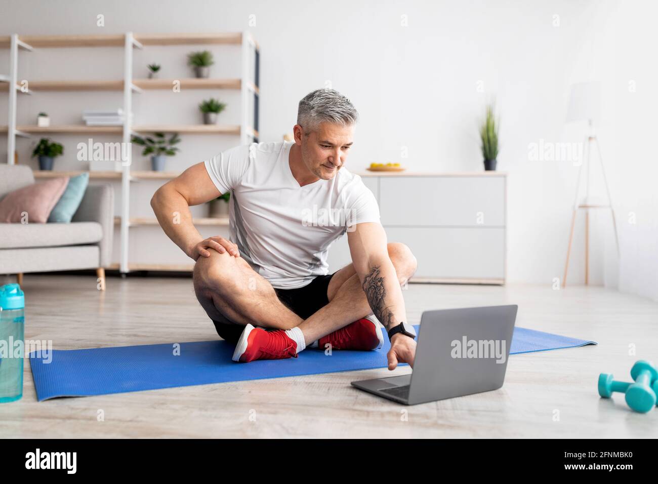 Uomo anziano che guarda il tutorial sportivo online sul computer portatile, esercitandosi a casa in salotto interno Foto Stock