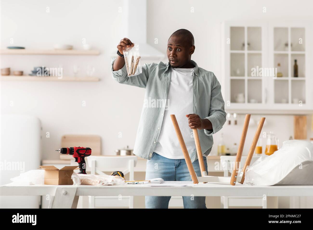 Rompicapo uomo afroamericano che tiene pacchetto con i dettagli, assemblando la sedia con le istruzioni in cucina Foto Stock
