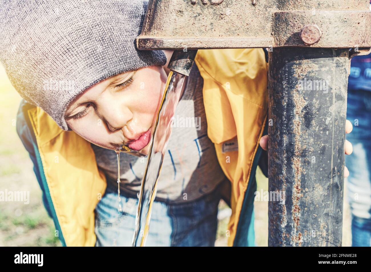 Divertente bambino piccolo in grigio cappello lavorato a maglia bevande acqua da pompa d'acqua d'epoca nel cortile della casa del villaggio il giorno di sole chiudi vista Foto Stock