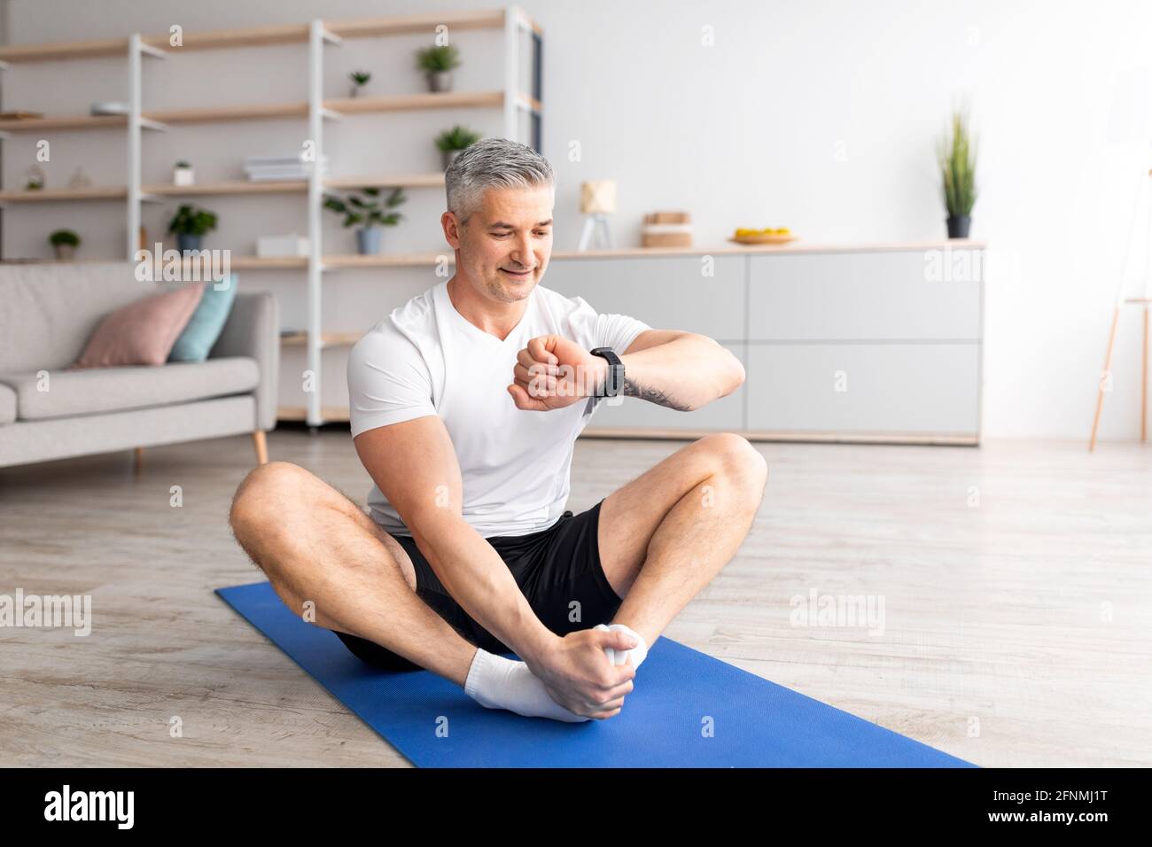 Tempo per la formazione. Uomo maturo che guarda il tracker di idoneità sulla sua mano, esercitandosi sul tappeto di sport a casa Foto Stock