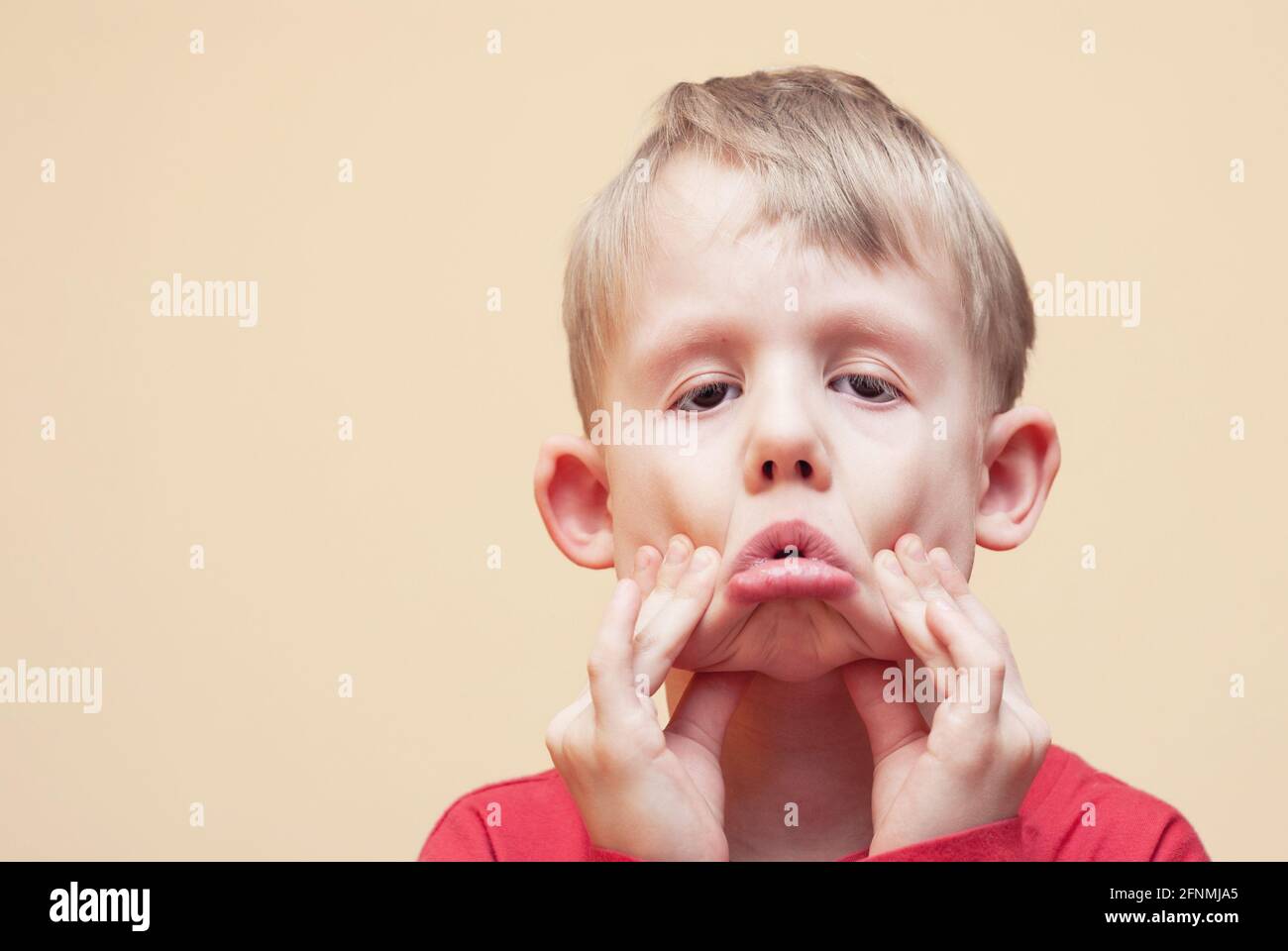 Ritratto di giocoso ragazzo biondo in abiti rossi con grimace posa per la fotocamera su sfondo beige in studio vicino visualizza Foto Stock
