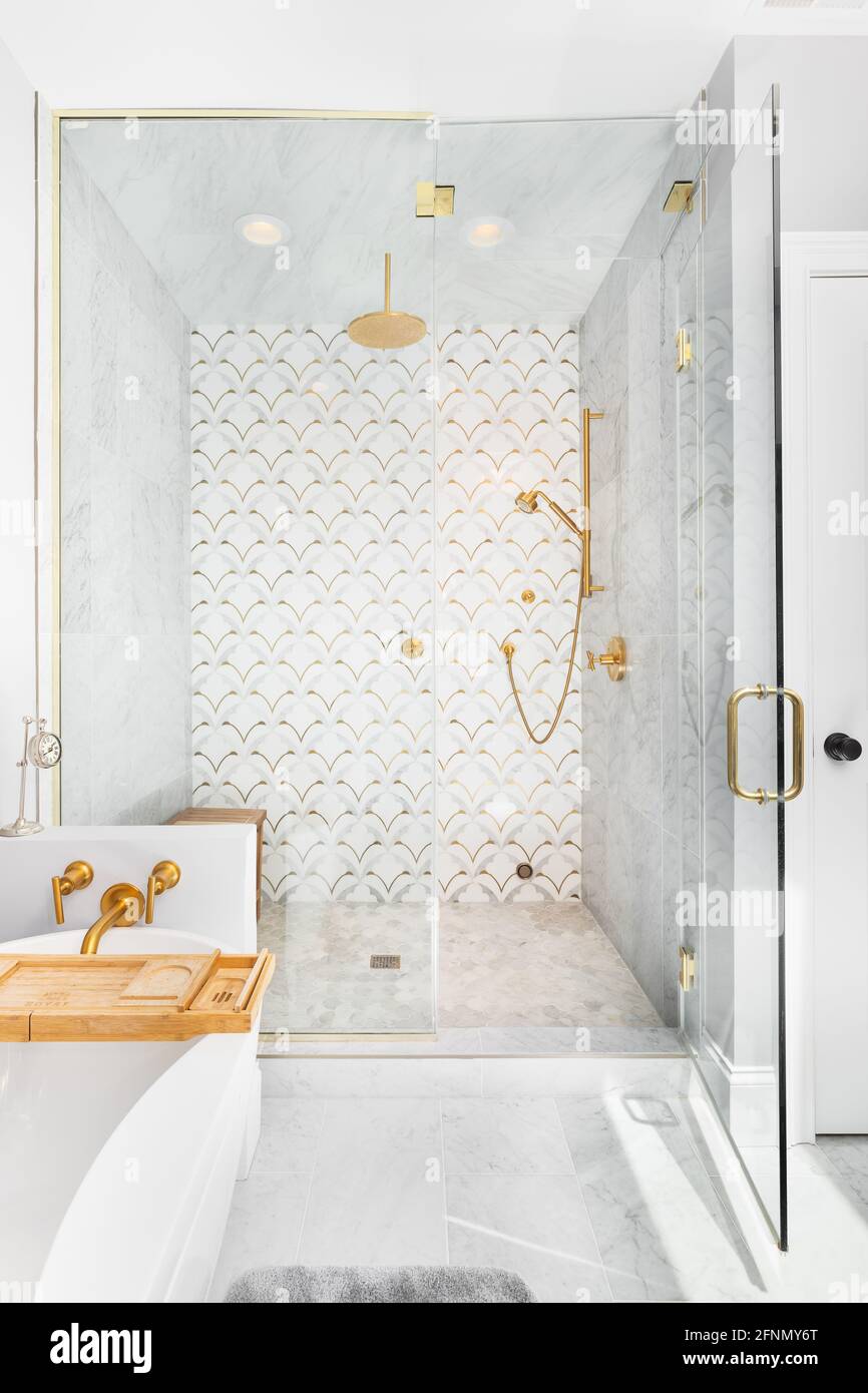 Una bella doccia e vasca piastrellata con piastrelle bianche e dorate sulle pareti, piastrelle di marmo mosaico sul pavimento, e soffione doccia e bulloneria dorati. Foto Stock