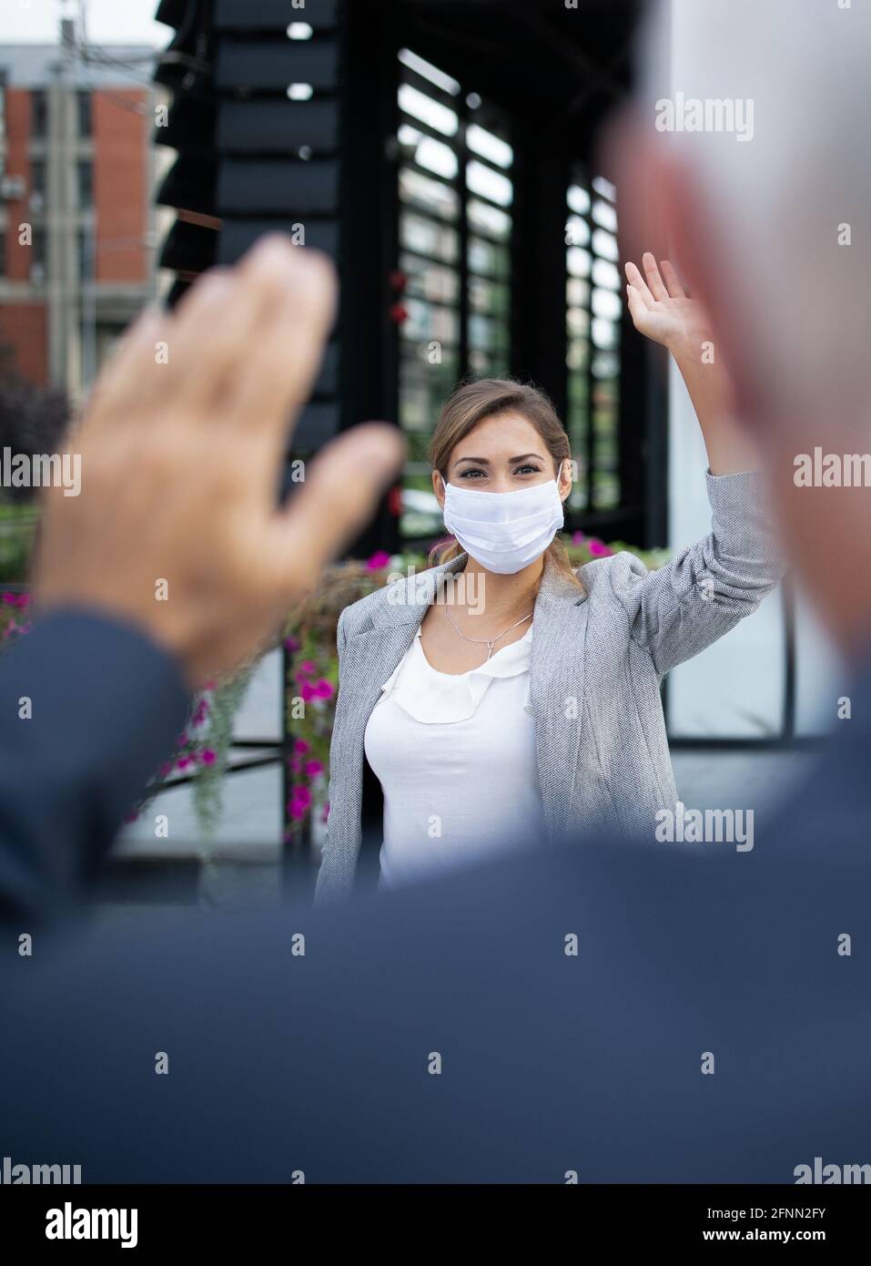 Donna d'affari con maschera di sicurezza e saluto dell'uomo agitando le mani di fronte all'edificio dell'ufficio. Protezione da virus e concetto di allontanamento sociale sul luogo di lavoro Foto Stock