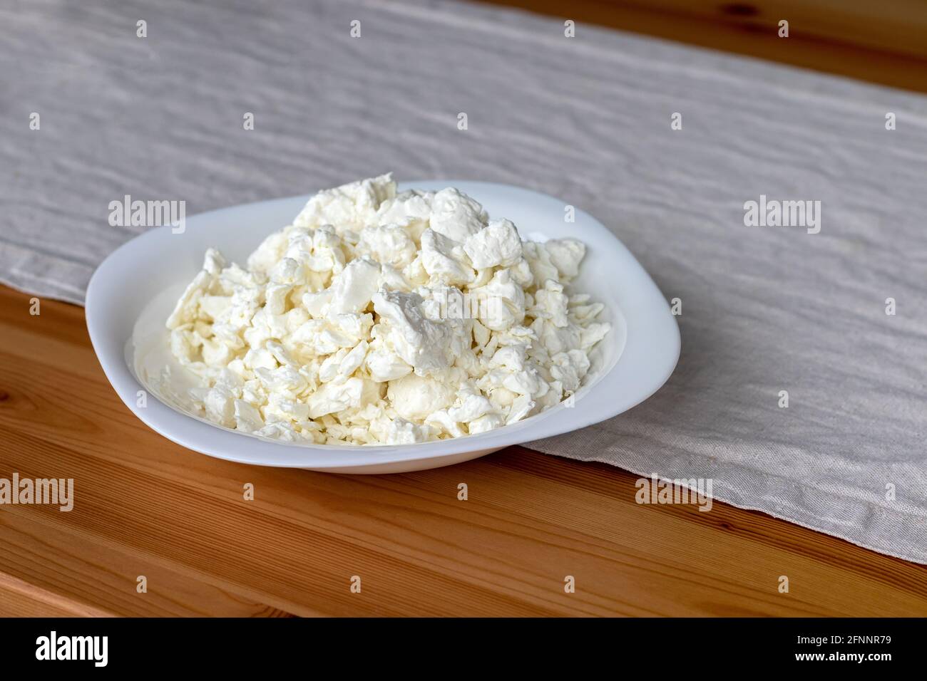 Un piatto bianco quadrato riempito di formaggio cottage si trova su una tovaglia di lino su un tavolo di legno. Messa a fuoco selettiva Foto Stock