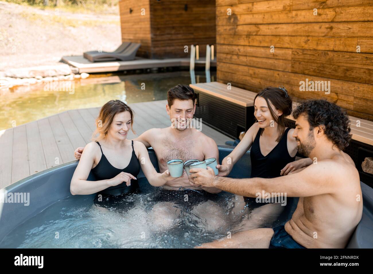 Compagnia di amici che fumano in un'iva all'aperto vicino al centro termale e lago. Donne e uomini si divertono e si rilassano nel centro benessere. Alzando gli occhiali e Grazie. Foto Stock