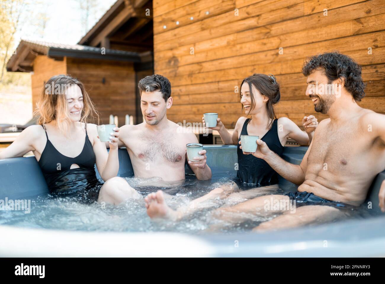 Compagnia di amici che fumano in un'iva all'aperto vicino al centro termale e lago. Donne e uomini si divertono e si rilassano nel centro benessere. Alzando gli occhiali e Grazie. Foto Stock