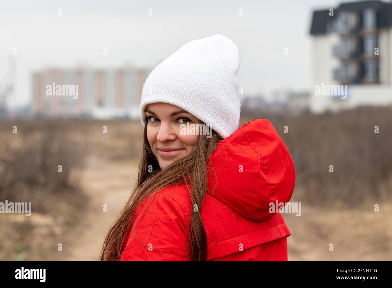 Giovane donna sorridente con capelli lunghi in cappello bianco lavorato a maglia e giacca rossa con cappuccio che guarda sopra la spalla la strada di giorno nuvoloso contro offuscata Foto Stock