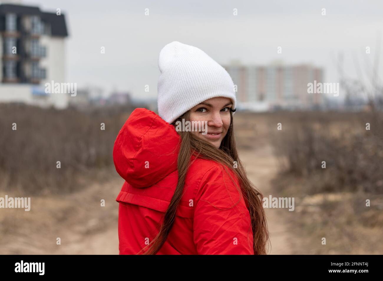 Giovane donna sorridente con capelli lunghi in cappello bianco lavorato a maglia e giacca rossa con cappuccio che guarda sopra la spalla la strada di giorno nuvoloso contro offuscata Foto Stock