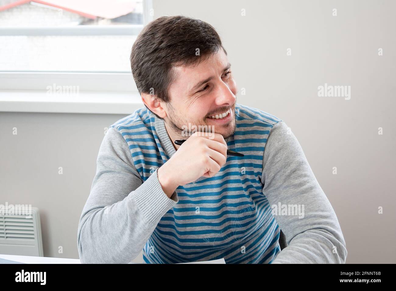 Bell'uomo sorridente in un maglione a righe si siede in un ufficio con una penna in mano e guardando via Foto Stock