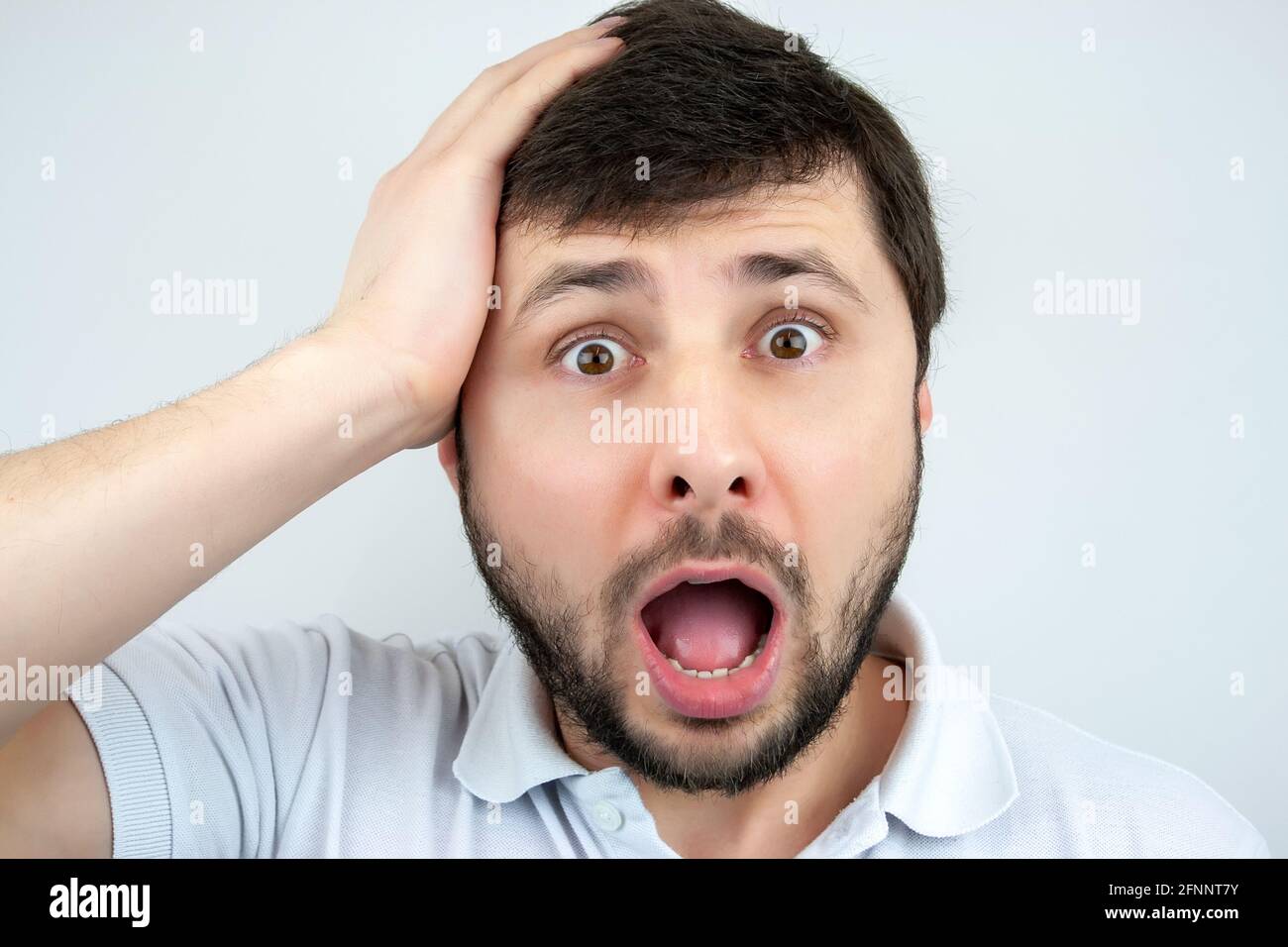 Ritratto di un uomo sorpreso bearded con occhi e bocca spalancati, tenendo la testa con la mano, in una T-shirt bianca su sfondo bianco Foto Stock