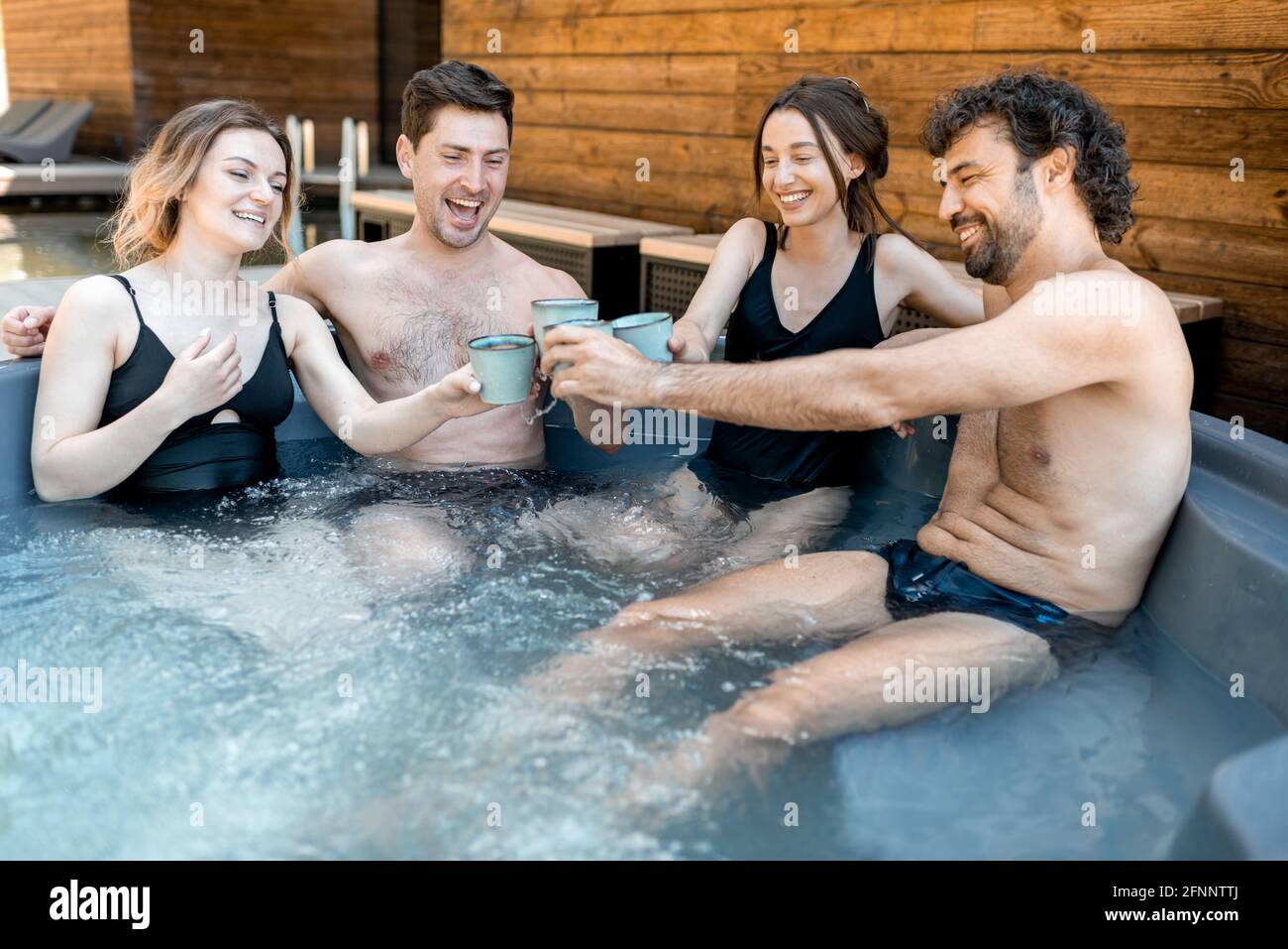 Compagnia di amici che fumano in un'iva all'aperto vicino al centro termale e lago. Donne e uomini si divertono e si rilassano nel centro benessere. Alzando gli occhiali e Grazie. Foto Stock