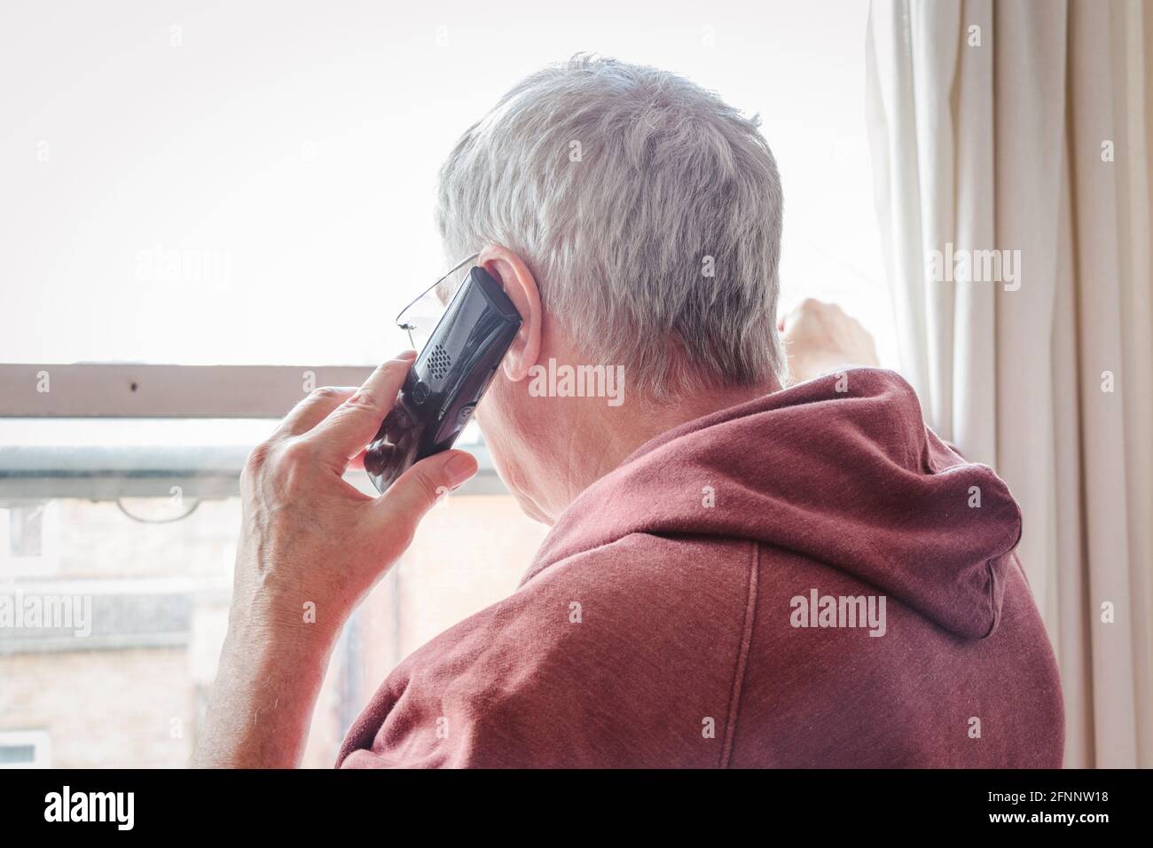 Vecchio uomo dai capelli grigi che usa il telefono che guarda fuori da una finestra, Londra, Regno Unito Foto Stock
