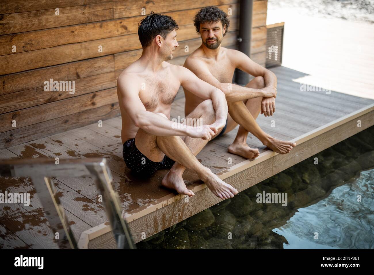 Amici maschi seduti sulla terrazza vicino al lago nel complesso termale e parlando. Rilassatevi e divertitevi nel centro benessere. Foto Stock